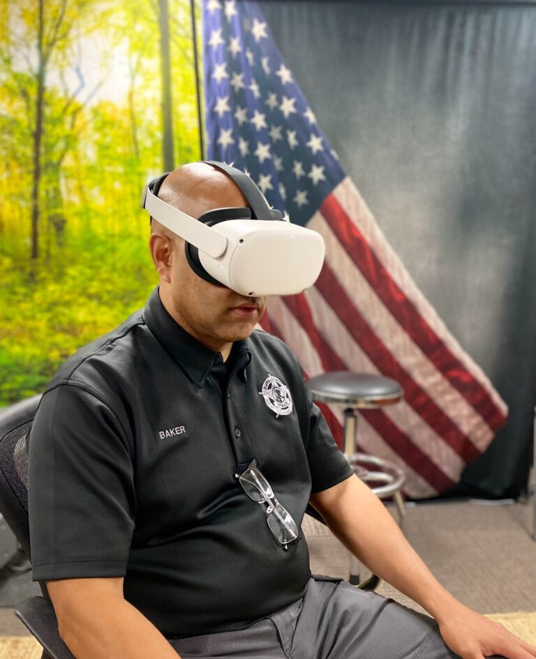 A man is sitting down in a wellness room. He is bald and wearing a black collared polo shirt with eyeglasses hanging from the middle. He is wearing a white virtual reality headset. The background is a forest scene on the left and a flag banner on the right.